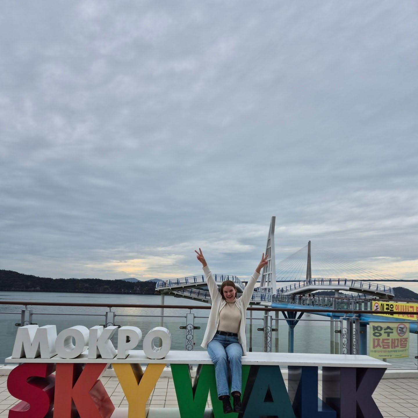 A traveller with both arms up, sitting on the MOKPO SKYWALK sign by the bridge