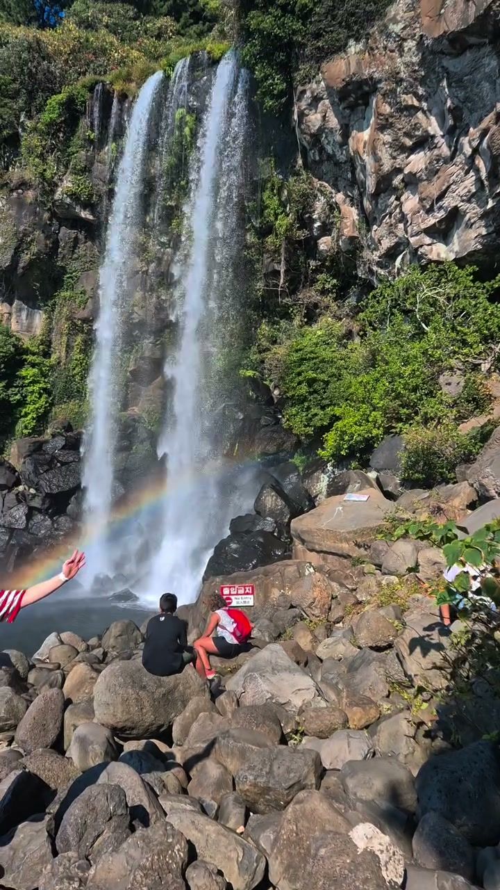 A tall Jeju waterfall over black volcanic rock, a rainbow in its spray